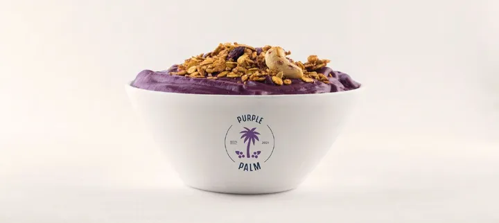 Selo circular da marca Purple Palm, com uma palmeira roxa, aplicado em uma tigela branca com creme roxo e granola