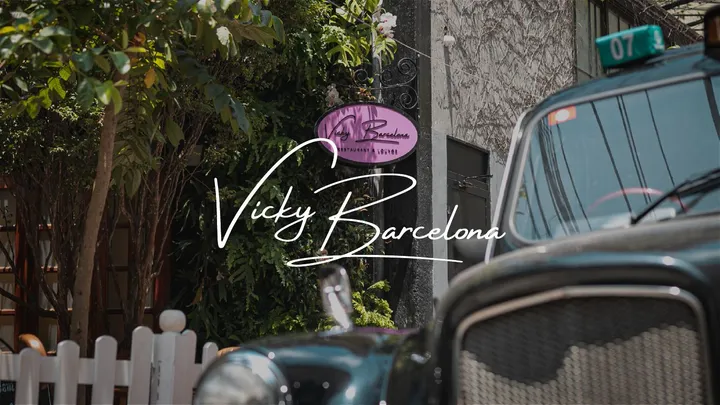 Fachada do restaurante com a placa rosa do Vicky Barcelona entre as folhagens e um carro antigo preto em primeiro plano, com o logotipo manuscrito Vicky Barcelona em branco sobre a foto